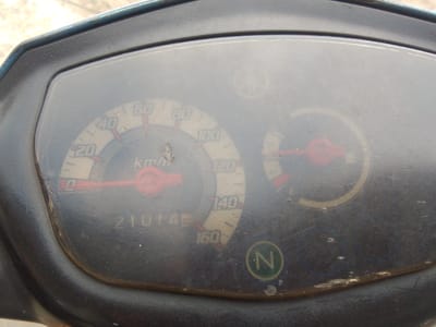 The image is a close-up of a motorcycle's instrument cluster. The cluster includes a speedometer with markings up to 160 km/h, and an odometer displaying "210145". There is also a fuel gauge and a neutral indicator light labeled "N". The instruments are covered by a slightly smudged and scratched plastic lens. The surrounding casing is black plastic. The background is blurred, but appears to be a light-colored, textured surface, possibly pavement. There are no people visible in the image. The image is taken from a slightly angled, overhead perspective. The overall condition of the instrument cluster suggests it is on a used motorcycle. This photograph was taken in Megamendung, Indonesia, and the scene is a motor vehicle.