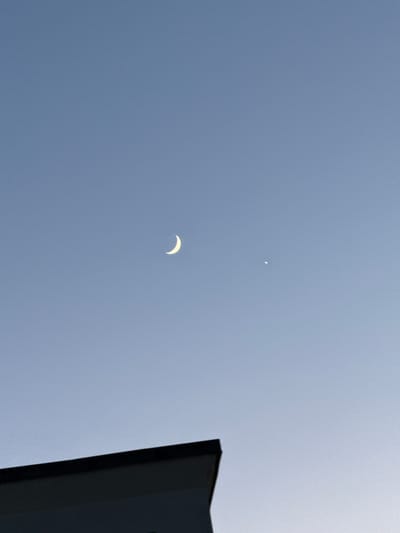 A crescent moon is visible in the clear, light blue sky over Glendale Heights, United States of America. To the right of the moon, a small, bright object, likely a planet, is also visible. In the foreground, the dark roofline of a building cuts across the bottom of the frame. The lighting suggests it is either early morning or late evening.