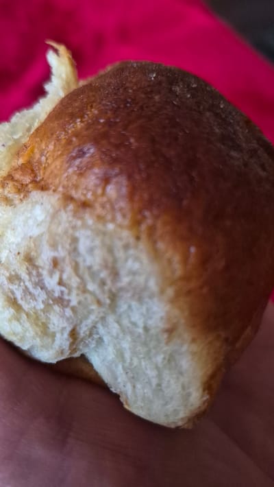 A close-up, slightly blurry shot shows a person's hand holding a piece of bread. The bread has a golden-brown, baked crust and a soft, white interior. The hand, with visible skin texture and some reddish tones, is positioned to cradle the bread. The background is out of focus, with a vibrant pink fabric to the left and a darker, reddish-brown area on the right, likely representing the rest of the person's clothing or another surface. There is no discernible text or specific time of day or weather indications. The image focuses on the texture and color of the bread and the human interaction with it.
