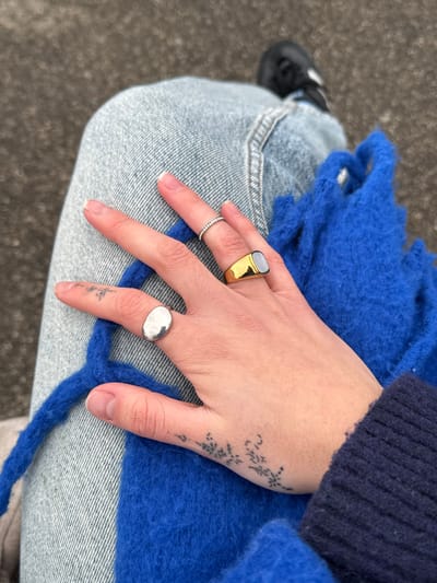 A person's hand adorned with three rings rests on their lap. The rings are on the index, middle, and pinky fingers. A gold ring with a gray stone is on the index finger. A slim ring with small, light-colored stones is on the middle finger. The pinky finger holds a round silver ring. A small black tattoo decorates the top of the hand. The person wears jeans and a blue sweater or scarf. Part of a black shoe is visible in the background.