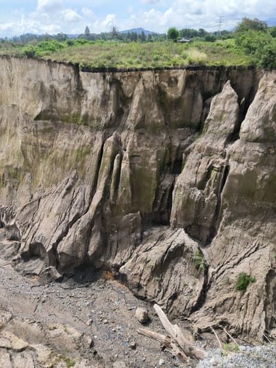 This media file depicts a dramatic landscape featuring a deep, eroded cliff face, likely a result of natural geological processes exacerbated by weather. The cliff is composed of layered soil and rock, showing signs of significant erosion with deep fissures, gullies, and overhangs. Patches of green vegetation, including grass and some shrubs, cling to the top edge of the cliff and sporadically grow on its sheer face, adding a touch of life to the rugged terrain. At the base of the cliff, a rocky, debris-strewn area is visible, with scattered pieces of wood and what appear to be fragments of plastic litter.

In the background, a lush green landscape stretches out, with trees and hills creating a natural horizon. In the distance, a small, simple building can be seen nestled among the greenery, and a prominent power pylon stands against the sky, suggesting proximity to infrastructure. The sky is partly cloudy with bright sunlight, indicating it is daytime. The overall scene evokes a sense of raw, untamed nature and the powerful forces of erosion. There is no visible text in the image. The location context of Kebunbaru, Indonesia, suggests a tropical or subtropical environment, which could explain the vibrant vegetation and the geological conditions leading to such erosion.