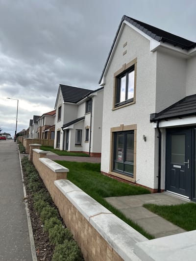 The image shows a row of newly constructed houses on a street in Dunfermline, United Kingdom. The houses are white with dark gray roofs and doors. Each house has a small patch of grass between the house and the sidewalk. A brick wall with a white concrete top runs along the sidewalk, separating the houses from the street. There are some shrubs and ground cover plants between the wall and the sidewalk. A streetlight is visible in the distance. Cars are parked along the street. The sky is cloudy. The houses appear to be located in a residential area on the outskirts of the city. A "SOLD" sign is visible in the window of one of the houses.