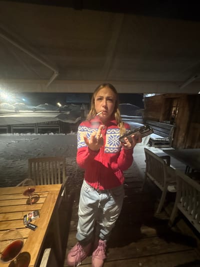 A young female stands outdoors on a wooden deck or terrace at night, in what appears to be a snowy environment, likely in La Clusaz, France. She has blonde hair and is wearing a red zippered cardigan with white and blue snowflake patterns, light blue ripped jeans, and pink boots. She holds a lit cigarette in her mouth, exhaling smoke, and extends the middle finger of her left hand towards the viewer. Her right hand holds a dark-colored smartphone, possibly an iPhone due to the camera bump visible on the rear. To her left, a wooden table is visible with two partially filled wine glasses containing red liquid, a pack of Marlboro cigarettes, and a small black lighter. The background reveals a snow-covered outdoor area with snow piled on structures and ground, illuminated by artificial light sources. A wooden building or chalet structure is visible further back on the right.