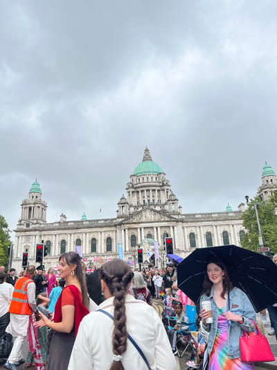 A wide-angle, eye-level shot captures a bustling scene in front of Belfast City Hall. The imposing neoclassical building, with its distinctive green dome and multiple towers, dominates the background under a cloudy sky. A crowd of people, dressed in a variety of attire including traditional and modern clothing, are gathered on the street. Many are holding small flags, some of which appear to be national flags and pride flags. A woman in the foreground to the right holds a black umbrella and a takeaway coffee cup, while another woman in the center foreground wears a red outfit and is looking towards the right. A man in an orange high-visibility vest is visible on the left. Traffic lights are illuminated red in the midground. The atmosphere appears to be festive, possibly during a parade or public event.