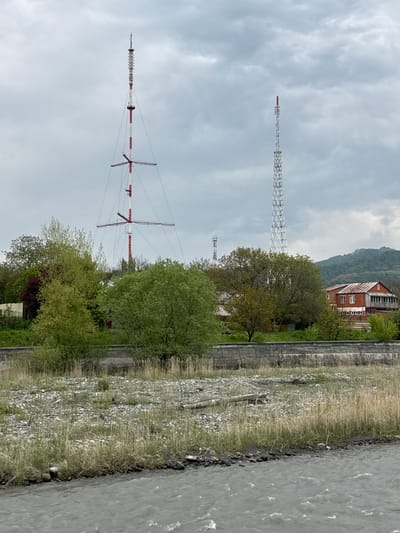 The image depicts a landscape in Assinovskaya, Russia, featuring two prominent radio towers. The foreground tower is painted with red and white stripes, while a more distant tower has a lattice structure. The towers rise above trees and vegetation. A building is situated on a hillside in the background. A riverbank with vegetation and a body of water occupy the right side of the frame. The sky is overcast.