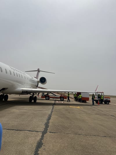 The image shows an airplane, likely a regional jet, parked on a concrete airport apron. Ground crew personnel wearing high-visibility vests are positioned near the aircraft, possibly performing maintenance or pre-flight checks. A ground support vehicle is also visible near the airplane. The location is possibly Kwi, Nigeria. The sky is overcast.