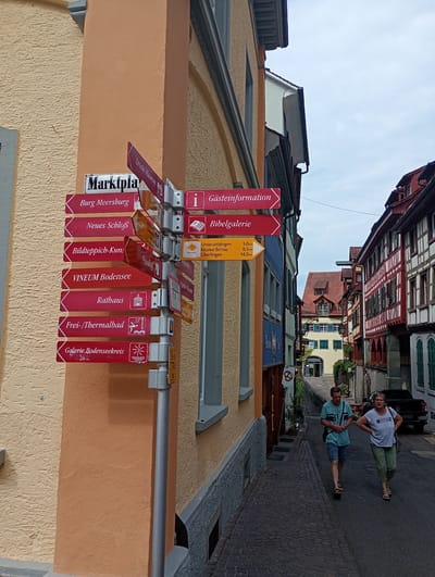 The image shows a narrow cobblestone street in Meersburg, Germany. On the left, a building with a textured orange facade is visible. A tall, silver metal pole stands on the right side of the street, adorned with multiple directional signs. These signs point to various locations, including "Marktplatz," "Burg Meersburg," "Neues Schloß," "Bildteppich-Kunst," "Vineum Bodensee," "Rathaus," "Frei-/Thermalbad," and "Galerie Bodenseekreis." Some signs also include distance markers in kilometers. On the right side of the street, there are half-timbered buildings with red and white facades. Two people are walking up the street, a man and a woman. The man is wearing a teal t-shirt and blue shorts, and the woman is wearing a white t-shirt with "Mustang" printed on it and green pants. The sky is overcast.