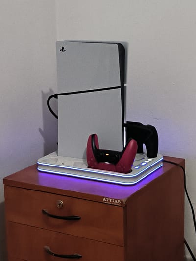 A PlayStation 5 console and two controllers are displayed on a white charging dock with blue LED lights. The charging dock is placed on top of a brown wooden desk with two drawers. A black cable is plugged into the back of the PlayStation 5 console. The desk is situated against a white wall. The scene appears to be indoors.