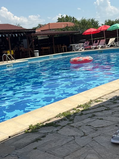 This media file shows a swimming pool area, likely at a resort or private residence, in Manolsko Konare, Bulgaria. The scene is oriented vertically, with the pool dominating the right side of the frame. The pool is filled with blue, patterned water, and a red and white inflatable ring is floating on the surface. The pool's edge is bordered by light-colored tiles and a stone patio with small green plants growing in the cracks.

On the left side of the frame, a covered seating area with wooden structures is visible. Several red and green umbrellas are extended, providing shade over lounge chairs. Behind the umbrellas and lounge chairs, trees and a blue sky with scattered clouds suggest it is a daytime scene with pleasant weather. The architecture of the covered area, with its tiled roof and wooden beams, provides a hint of the location's style.

In the distance, what appears to be a building with a red-tiled roof can be seen among the trees. The overall atmosphere is one of relaxation and leisure, typical of a sunny day spent by a pool. No people are clearly visible, but the presence of lounge chairs, umbrellas, and the inflatable ring indicates that the area is set up for human activity. The time of day appears to be late morning or early afternoon, given the lighting and the clarity of the sky. No visible text is present in the media file.