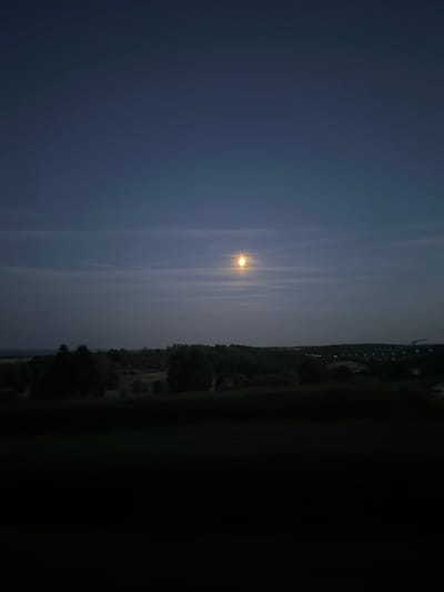 The image depicts a landscape view at dusk or night, featuring a full moon partially obscured by thin clouds. The moon is positioned in the upper portion of the frame, casting a soft light over the scene. Below the sky, a dark silhouette of rolling hills and trees stretches across the horizon. In the distance, faint lights suggest the presence of a town or infrastructure, possibly a bridge or construction site, indicated by a visible crane. The foreground is dominated by a dark, indistinct area, likely a field or hill. The overall ambiance is serene and twilight, characteristic of a rural or semi-rural environment. The location is identified as Méan, Belgium.