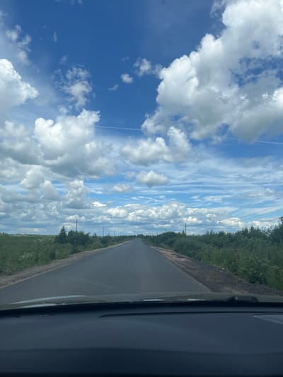 This is a vertical, low-angle, first-person perspective shot of a road in Губачево, Russia. The road stretches into the distance, bordered by lush green trees on the left and what appears to be a dark, greyish material on the right, possibly gravel or a dark soil. The sky is a dominant feature, filled with a dramatic mix of white, puffy cumulus clouds against a vibrant blue expanse. Wisps of cirrus clouds are also visible, creating a textured, layered effect. A faint contrail from an aircraft is present in the upper left quadrant of the sky. The perspective suggests the image was taken from a vehicle, possibly from the passenger seat looking out the window. The lighting and bright clouds indicate it is daytime, likely late morning or early afternoon. There are utility poles with wires visible along the right side of the road, suggesting it is a rural or semi-rural area. No people are visible in the frame. The overall mood is one of open space and natural beauty, with the dynamic sky drawing significant attention.