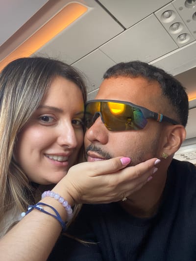 Two people are in close proximity, posing for a selfie inside an airplane cabin. The woman, on the left, has long, blonde-streaked brown hair and is smiling broadly, her left hand gently touching the man's cheek. She is wearing two beaded bracelets on her left wrist, one purple and white, the other dark blue with white beads. The man, on the right, is wearing sunglasses with orange-tinted lenses that reflect the cabin's interior and a portion of the woman's face. He has short, dark hair and is wearing a dark shirt. A small gold earring is visible on his right earlobe. The overhead compartment and lighting of the airplane are visible in the background. The overall lighting suggests the photo was taken during daytime or with artificial cabin lighting.