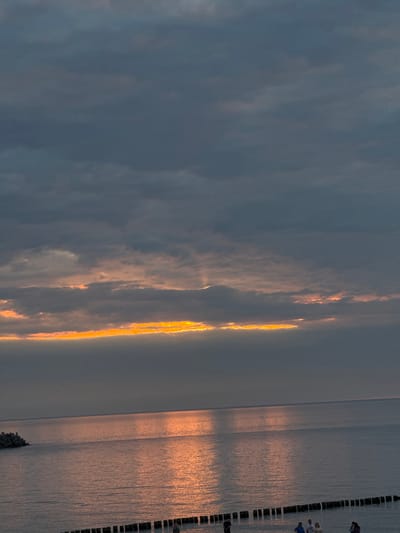 This image captures a serene sunset over the Baltic Sea at the port of Ustka, Poland. The sky is a dramatic canvas of dark, moody clouds, pierced by a vibrant band of golden-orange light where the sun is setting just below the horizon. This warm glow reflects beautifully on the calm water, creating shimmering streaks of light.

In the foreground, a line of wooden breakwaters extends into the sea, with a few scattered people visible near them. To the left, a section of the Ustka pier or breakwater structure juts out into the water. The overall atmosphere is one of tranquility and natural beauty as day transitions into evening.