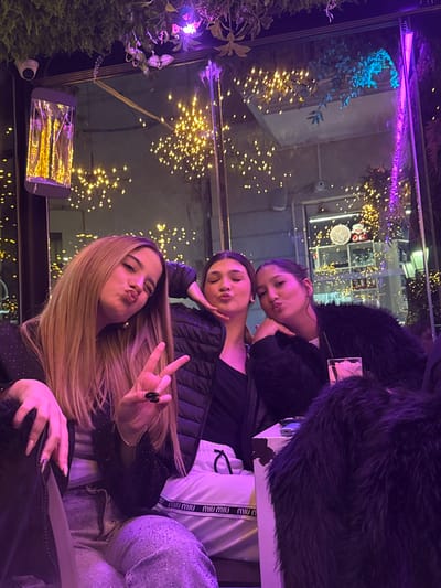 This image captures three females seated indoors, likely at a bar or cafe, in Stornara, Italy, during nighttime. The setting features prominent purple ambient lighting and numerous warm yellow string lights, both actual and reflected in a large window or mirror behind the subjects.

From left to right:
The first female has long, light brown hair. She is wearing a dark, possibly embellished jacket and light-colored, sparkling pants. Her left hand displays a "peace" sign with black-painted fingernails, while her lips are pursed in a "duck face."
The second female, with dark hair pulled back, is wearing a black puffer jacket over a dark top and white sweatpants with "Miu Miu" branding on the waistband. Her right hand is positioned near her chin, and her lips are pursed in a "duck face."
The third female, also with dark hair pulled back, is wearing a black faux fur jacket. Her right hand is positioned near her chin, and her lips are pursed in a "duck face."

On a white surface, likely a table, in front of the rightmost female, a clear glass containing a beverage with ice is visible. The background shows reflections of numerous small, scattered lights. Above the subjects, decorative foliage with purple lighting is visible, along with a wall-mounted rectangular light fixture and a small security camera on the upper left. Through the reflective surface, an entrance or doorway with additional lighting and a circular illuminated sign is discernible further in the background.
