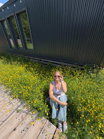 An overhead shot, rotated 90 degrees clockwise, captures a young woman relaxing outdoors amidst a vibrant field of yellow flowers in Vozhoiskii, Russia. She is lying on her back, her head positioned towards the upper right of the frame, facing the camera with a relaxed smile. Her right hand is raised, adjusting a pair of sunglasses perched on her nose.

The woman is dressed in a white sleeveless crop top, light blue jeans, and white sneakers with patterns. A watch is visible on her left wrist, and she wears a simple necklace. She appears happy and at ease in the bright sunshine.

Surrounding her is a dense expanse of small yellow flowers, likely buttercups, interspersed with green foliage. These flowers cover much of the ground, creating a cheerful, natural carpet. To the left side of the frame, a modern building with dark grey corrugated metal siding runs vertically. Several horizontal rectangular windows are visible on the building, reflecting green trees or vegetation. Along the bottom right, a wooden deck or pathway with light-colored planks contrasts with the greenery and flowers. Strong sunlight is evident, casting distinct shadows of the flowers onto the wooden surface. The scene suggests a pleasant, sunny day. No visible text is present in the image.
