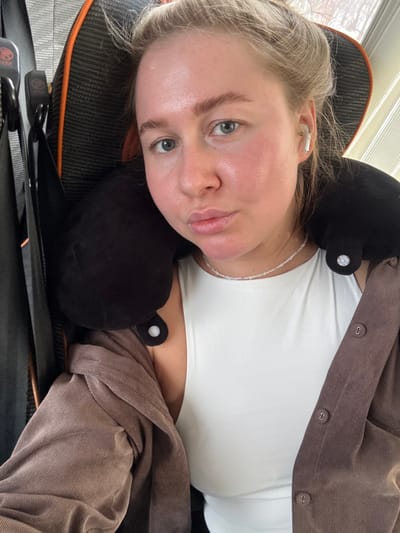 In the city of Гремяча, Russia, a fair-skinned female individual is depicted indoors, seated. Her blonde hair is pulled back, revealing light-colored eyes and full, pursed lips. She wears a white sleeveless top, an unbuttoned light-brown corduroy-textured shirt, and a thin, light-colored beaded necklace. A black travel neck pillow, secured by white snap fasteners, is positioned around her neck. A white wireless earbud is visible in her right ear. She is seated in a black and orange gaming or office chair, which features a visible headrest and a partially obscured logo. The background contains vertical window blinds, indicating an indoor environment.