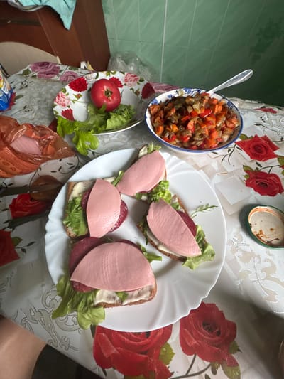 A top-down shot shows a meal laid out on a table covered with a floral tablecloth in Krasnoye, Russia. The table is set with a white plate of four open-faced sandwiches, each topped with lettuce, a slice of red sausage, and a thick slice of pink bologna. To the right of the sandwiches is a small green jar with a metal lid, and further to the right, a bowl of stewed vegetables including peppers, carrots, and onions. Behind the sandwiches, a white plate holds a single red tomato and a small pile of lettuce leaves. A bag of sliced bologna is partially visible on the left side of the table. The background shows a green tiled wall and wooden cabinets.