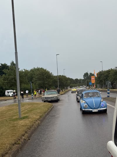 The image depicts a street scene in Falkenberg, Sweden, on a cloudy day, with wet asphalt indicating recent rain. A procession of vintage cars is moving along the road. In the foreground, a blue Volkswagen Beetle with license plate "DGT 109" is prominent, followed by other classic cars, including a yellow one. In the background, a light-colored older car is also visible. On the left side of the road, several people are standing and walking, some on bicycles. A white van with "RESCUE" written on it is parked on the side. Trees line the background. The overall atmosphere suggests a car rally or event.
