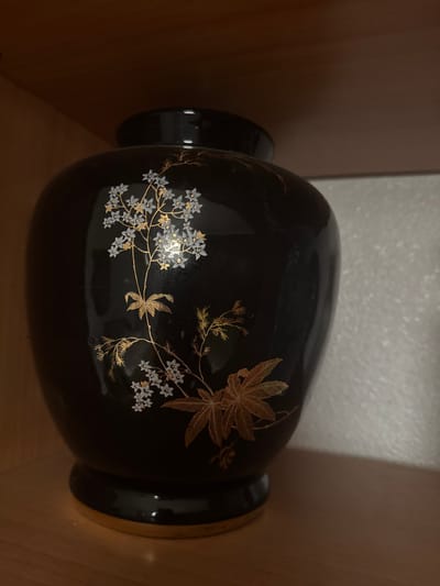 A black, glossy vase with gold and white floral motifs sits on a wooden shelf in a room. The vase is bulbous in shape with a slightly narrowed neck and a flared base. The floral design features delicate white flowers with gold accents, and larger, feathery gold leaves. The background is a textured white wall on the right and a wooden surface on the left, suggesting a shelf or cabinet. The lighting is dim, with highlights reflecting off the vase's surface. There are no people or animals present in the image. The scene is indoors and appears to be a still life.