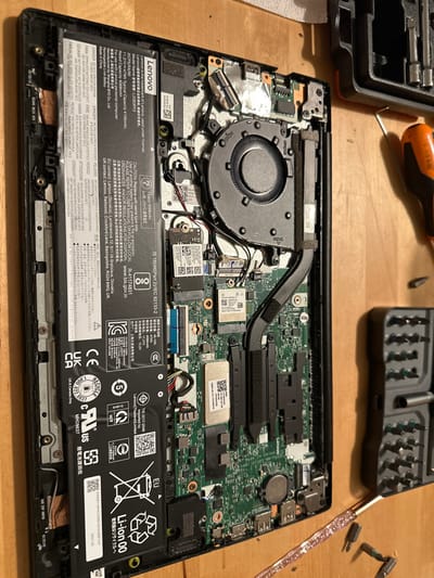 The image shows the internal components of a Lenovo laptop, particularly focusing on the battery, cooling fan, and motherboard. The black Lenovo battery occupies a significant portion of the lower section, displaying warnings and safety information in multiple languages. A cooling fan is positioned near the top left, connected to a heat pipe. Various chips, connectors, and other electronic components are visible on the motherboard, showcasing the intricate design of the laptop's internal circuitry. Several small screwdrivers and a case with various driver bits are visible.