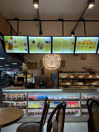 The image depicts the interior of a cafe in Mai Khao, Thailand, likely an airport cafe due to the background and overhead lighting. In the foreground, two chairs with woven backs are visible. The cafe counter displays a variety of pastries under glass, with prices indicated. Behind the counter, shelves are stocked with packaged food items such as instant noodles, snacks, and drinks.
Above the counter, three digital menu boards are illuminated. The leftmost board advertises "RICE DISHES" with images of food and prices. The middle board features "PHUKET LOCAL FLAVORS" and "PERFECT MEAL DUO" with food and drink options and prices. The rightmost board shows "CLASSIC COMBO SET" options including pastries and drinks.
A prominent circular neon sign above the counter reads "LE CORAL CAFE - ALWAYS FRESH" with a sun graphic. Another sign next to it says "KEEP CALM AND DRINK COFFEE". An analog clock with Roman numerals is mounted on the tiled wall behind the counter.
Several people are visible in the background, appearing to be customers and staff. Luggage is present, further suggesting a travel hub. The overall environment appears clean and well-lit, with a modern design.
