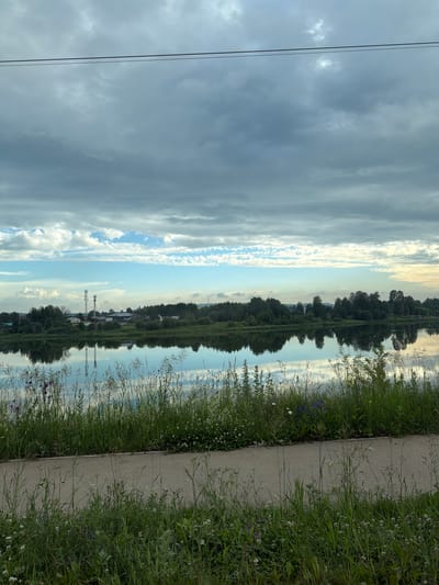 The image depicts a tranquil waterscape with a reflection of the sky and a distant treeline. The foreground features a paved pathway running parallel to the water, bordered by lush green grass and wild flowers. The sky is a mix of grey and white clouds, with patches of blue visible. The water is calm and reflects the scenery, creating a mirror image. A settlement with buildings and communication towers is visible on the opposite bank of the water. The overall impression is one of natural beauty and serenity. The location is the city of Yug, Russia.