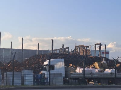 The image depicts the aftermath of a fire at an industrial site in Plastik, Russia. The foreground shows a flatbed truck laden with palletized goods, possibly building materials, covered in protective white plastic sheeting. In front of the truck, stacks of what appear to be metal cages or wire mesh containers are visible. Beyond these, a large pile of burnt debris, including charred wood and twisted metal, dominates the scene, suggesting significant structural damage. In the background, the skeletal remains of a large building are visible, with exposed framework and partially collapsed sections. A white building with red rectangular accents stands to the right, seemingly unaffected by the fire. The sky is clear blue with scattered white clouds. The overall impression is one of destruction and recovery.
