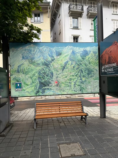 A digital billboard displays a detailed topographical map of the Mont-Blanc mountain range, specifically highlighting the ski resort of Saint-Gervais-les-Bains, France. The map is large, spanning the width of the frame, and features intricate details of slopes, trails, and elevations. A red marker is visible on the map, indicating a specific point of interest. To the right of the map, a smaller advertisement for "Émotions ALPİNES" with a website address is displayed. In the foreground, a wooden park bench with a dark metal frame sits on a tiled pedestrian area. The ground is paved with dark grey square tiles, and a metal grate is visible in the lower right corner. Behind the bench and billboard, buildings with traditional European architecture are visible, including one with white shutters and balconies. A tree with lush green foliage is in the upper left corner of the image. The scene appears to be an outdoor public space, likely a bus stop or information point, in the town of Saint-Gervais-les-Bains.