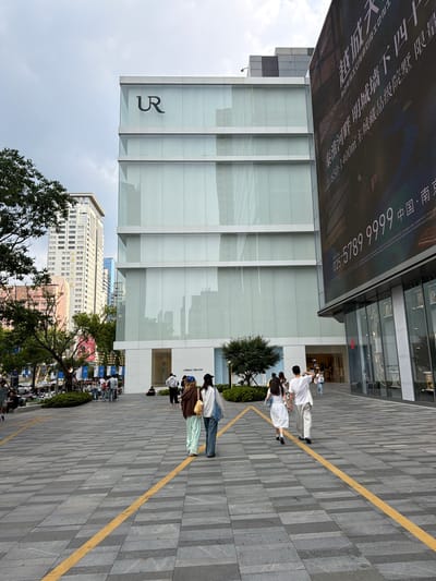 The image shows a street scene in the Qinhuai District of China, with a focus on a modern, multi-story building with a glass facade. The building displays the "UR" logo prominently at the top. To the right, a large digital billboard displays text in Chinese, along with a phone number. In the foreground, a paved walkway with yellow directional lines leads towards the buildings. Several people are walking on the walkway. The environment suggests an urban commercial area with pedestrian traffic. Trees are visible on the left side of the image, and other buildings are in the background, indicating a city setting.