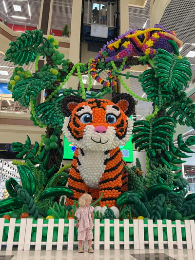 This image shows a balloon sculpture of a tiger in what appears to be a shopping mall or indoor public space. The tiger is the central focus, made entirely of balloons in orange, black, and white. It is surrounded by balloon sculptures of jungle foliage, including trees and large leaves in various shades of green. Above the tiger, a colorful balloon sculpture of a chameleon hangs. In the foreground, a young girl with blonde hair, wearing a pink patterned outfit, stands behind a white picket fence, looking towards the right side of the frame. The environment features a tiled floor and a background that includes structural elements of a large building, possibly an elevator shaft with people visible inside. The setting does not suggest the city of Gaponovskii, Russia, or any specific city.