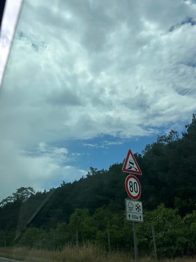 The image depicts a rural mountainous scene in Gorno Varshilo, Bulgaria, viewed from inside a vehicle, evidenced by a glare on the left. The upper two-thirds of the frame features a partly cloudy sky with patches of blue visible amidst greyish-white clouds. Below the sky, a dense, dark green forested hillside slopes from left to right across the mid-ground. A tall, multi-component road sign stands on the right side, against the backdrop of the forest. The topmost sign is a red-bordered white triangle with a black skidding car symbol, indicating a slippery road. Below it is a red-bordered white circular sign displaying "80" in black, signifying a speed limit of 80. The bottom sign is a white rectangle with symbols for rain and snow, and the text "↑ 600M ↑", likely indicating a distance for the depicted conditions. The immediate foreground shows dry, light brown grass and sparse green vegetation along what appears to be a roadside. No individuals or active subjects are visible.