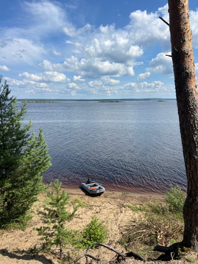 An inflatable boat with an outboard motor is beached on a sandy shore beside a wide river under a partly cloudy sky. The foreground features sand, small pine trees, and the trunk of a large pine tree on the right. The middle ground is dominated by the dark blue water of the river, with ripples indicating a slight breeze. In the distance, the opposite shore is visible, lined with trees and a hint of distant industrial structures. The sky is bright blue with scattered white cumulus clouds. The scene appears to be located in a natural, undeveloped area. This scene could be found on the Oka River in the city of Orel, Russia, which is known for its scenic riverbanks and pine forests.