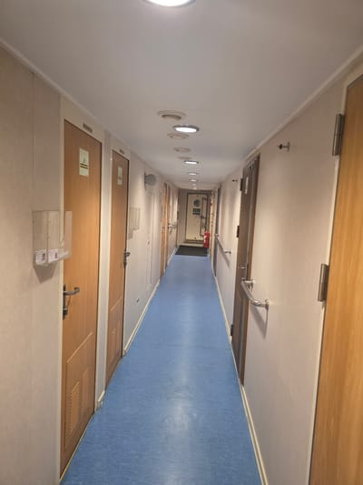 The image depicts a long, narrow corridor with doors on either side. The corridor has a blue floor and a white ceiling with circular lights. The walls are a light beige color. On the left side, there are several wooden doors with metal handles and signs above them. There are also clear plastic holders with what appear to be identification cards attached to the wall next to some doors. On the right side, there are also wooden doors and metal handrails along the wall. Towards the end of the corridor, there is a door with an "EXIT" sign and a red fire extinguisher next to it. The overall impression is of a functional interior space, possibly on a ship or in a building with many rooms. There is no information in the image that would suggest a specific location in Brindisi, Italy.