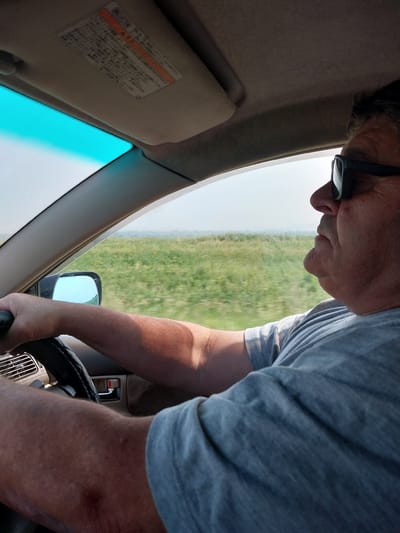 A medium shot captures a person driving a car, with the camera positioned inside the vehicle and looking forward. The driver, wearing a grey t-shirt and sunglasses, is holding the steering wheel with their right hand. Their left arm is visible resting on the steering wheel. The interior of the car is visible, including the dashboard, a side mirror, and a sun visor with Japanese text. Outside the car, a blurry green landscape with a hazy sky can be seen through the windshield and side window. The image does not provide any specific details about the city of Sychevka, Russia.
