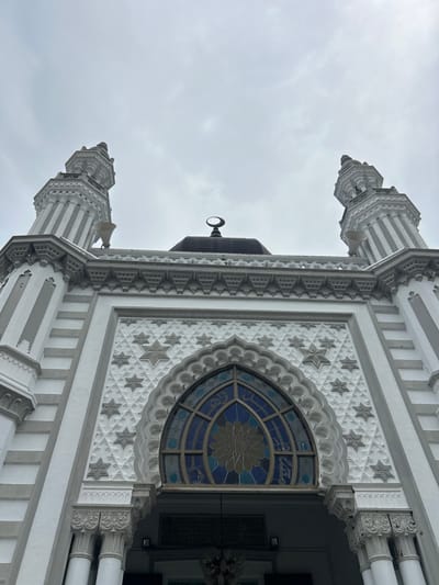 The image shows a low-angle, full-frame shot of the Zahir Mosque in Alor Setar, Kedah, Malaysia. The mosque's facade is white with intricate grey geometric patterns and star motifs. Two tall, slender minarets flank the central structure, topped with decorative spires. Above the main entrance, a large, arched stained-glass window features a floral design in shades of blue and yellow. A crescent moon and star emblem is visible on top of the dome, centered between the minarets. The sky is overcast with a dull, grey cloud cover. No people are visible in the image.