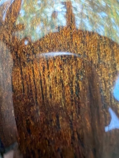 The image displays a close-up, abstract view of a textured surface. The surface has a predominantly brown and golden hue with vertical striations and a rough, almost granular texture. There are also areas of lighter, reflective patches, suggesting a glossy or wet finish. The background is blurred and appears to be a reflection of the surroundings, with hints of green, blue, and white light. The specific location within Southlake, United States of America, is not discernible from the visual information provided.