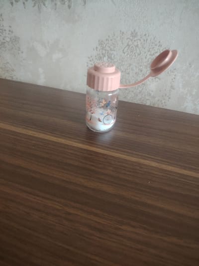 A clear glass bottle with a pink lid and flap is centered on a brown wooden table. The bottle contains a white powdery substance. The bottle has a design of butterflies and a bicycle wheel. The background is a beige wall with a floral pattern. The image appears to be taken indoors. The location of Hovsan, Azerbaijan is not discernible from the image.