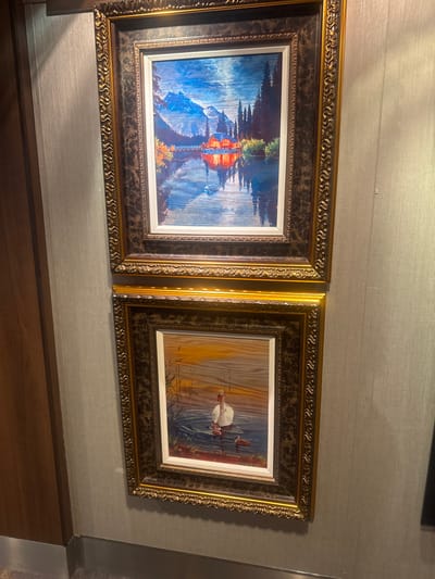 Two framed paintings hang on a textured wall. The top painting depicts a mountainous landscape with a body of water in the foreground, reflecting a cabin and trees illuminated by warm light. The bottom painting shows a swan and cygnets on water, with reeds in the background. Both frames are ornate with gold and dark brown accents. To the left of the paintings is a wooden door frame. The location in the city of Motril, Spain is not discernible from the image.