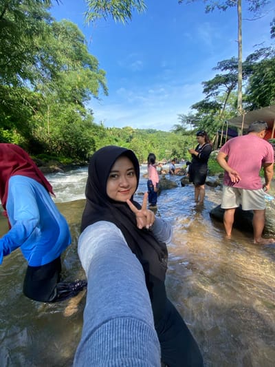 A group of people are enjoying time at a waterfall. One person, wearing a black hijab and gray sweater, is taking a selfie, holding up a peace sign. Others are wading in the shallow water behind them. The surroundings are lush with greenery and a clear blue sky is overhead. The water appears shallow and flowing. Some people are fully clothed.