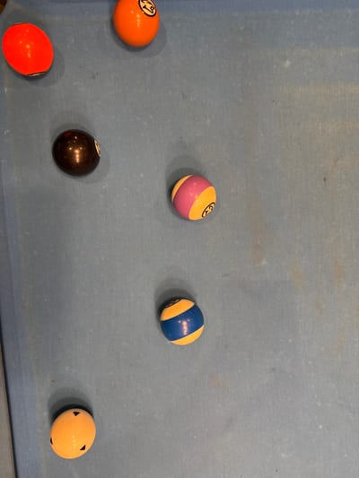 The image depicts a blue pool table with six pool balls scattered on the surface. The balls are: an orange 13-ball, a purple/yellow striped ball, a blue/yellow striped ball, a black 8-ball, an orange ball, and a red ball. The pool table shows signs of wear. The location in Bellville, South Africa is unknown.