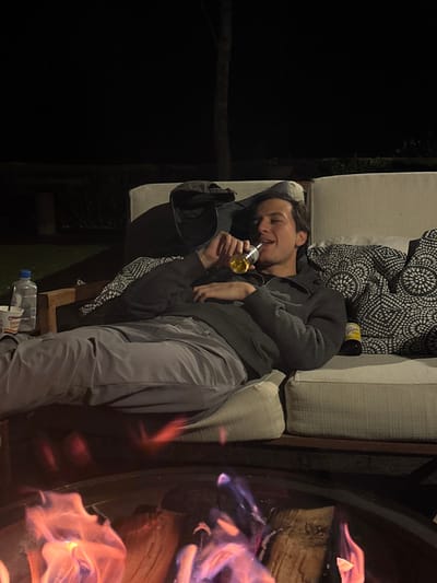 The image displays an outdoor nighttime scene in Huasca de Ocampo, Mexico. A male individual, appearing to be an adult, is reclined on a light-colored outdoor sectional sofa. He wears a dark grey hooded top and light grey trousers. His head rests on a pillow with a black and white geometric pattern. He holds an amber-colored bottle, identified as a Corona beer bottle, to his mouth, actively drinking from it. His eyes are partially closed, and his mouth is slightly open. Another Corona beer bottle rests on the sofa to his right. To his left, on an adjacent surface, a plastic water bottle with a blue cap and a white paper cup labeled "Maruchan Instant Lunch" are visible. A dark-colored baseball cap is situated on the sofa near his left shoulder. In the foreground, a circular metal fire pit contains burning logs, emitting bright orange, red, and purple flames. The background is dark, with vague silhouettes of trees or vegetation.