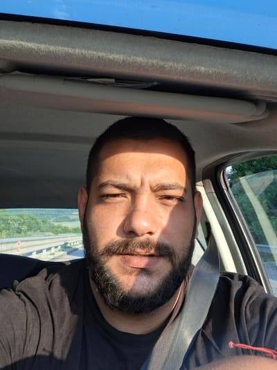A close-up, selfie-style image shows a man with dark hair, a beard, and a black t-shirt, seated in the driver's seat of a vehicle. He is wearing a seatbelt. The background reveals a highway and a landscape of trees and fields, suggesting a rural or suburban setting outside of a city. The lighting indicates it is daytime, with sunlight casting shadows on the man's face and shirt. The image was taken in or around Rashkovo, Bulgaria.