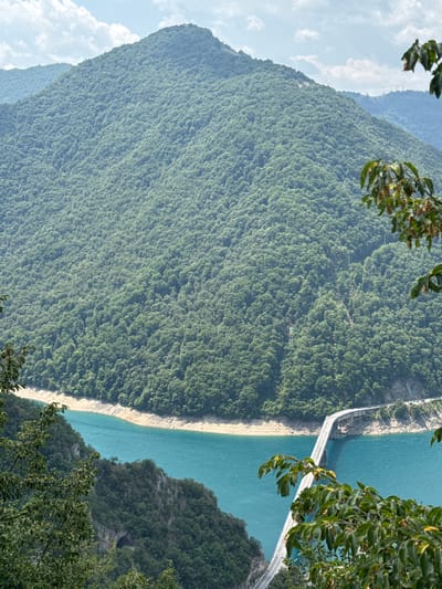 This image depicts a picturesque landscape scene near Unač, Montenegro, dominated by a vast, tree-covered mountain rising majestically. A strikingly blue body of water, likely a lake or river, carves through the base of the mountain, its shores marked by a sandy embankment. Spanning the water is a long, modern bridge, its white structure contrasting sharply with the surrounding natural elements. The bridge appears to carry a road, with vehicles subtly visible in the distance on its far end. The foreground is adorned with lush green foliage, framing the view of the mountain and water. The sky above is a soft blue, dotted with scattered white clouds, suggesting a clear, pleasant day. The overall impression is one of serene natural beauty and a harmonious blend of infrastructure with the environment.