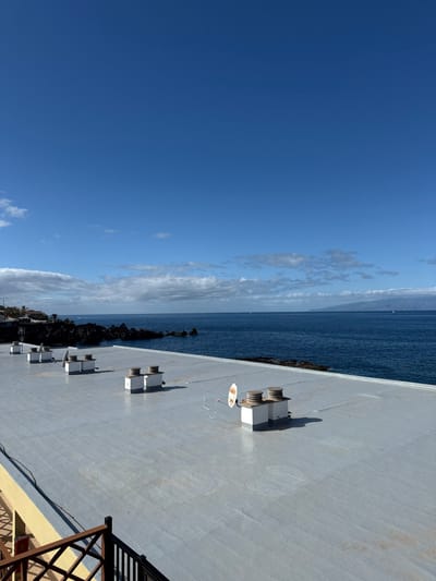 The image displays a panoramic view from a flat, light gray rooftop located in Santiago del Teide, Spain, overlooking the Atlantic Ocean. In the immediate foreground, the bottom-left corner reveals a portion of a yellow-painted building and a dark brown wooden railing. The rooftop surface exhibits five white, block-like ventilation units, each topped with a dark circular cap. One unit on the right side of the rooftop has a small, white, circular satellite dish attached, with white cables extending from it onto the roof. The rooftop surface shows minor staining and discoloration. Shadows cast by the structures suggest direct sunlight from a high angle.
Beyond the rooftop, the dark blue expanse of the Atlantic Ocean is visible, extending to the horizon. To the mid-left, a rugged, dark volcanic rock coastline juts into the sea. Several small white sailboats are discernible on the distant water. On the far right horizon, a large landmass, presumably an island with mountainous terrain, is visible under a slight haze. The sky is predominantly clear and bright blue, with scattered white cumulus clouds concentrated along the horizon and extending slightly into the upper left. No individuals are present in the scene.