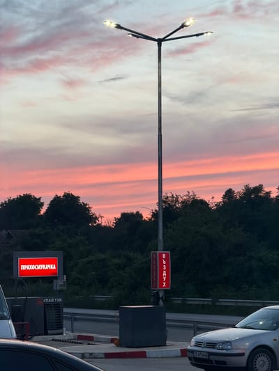 The image depicts a gas station in Dalgopol, Bulgaria, during sunset. The sky is filled with vibrant pink and orange hues, a result of the setting sun filtering through the clouds. In the foreground, a silver Volkswagen Golf is parked, its license plate partially visible. To the left of the car, a gas pump is partially visible, along with a red sign displaying text in Cyrillic: "ПРАХОСМУКАЧКА" which translates to "vacuum cleaner". A tall street lamp with multiple lights stands prominently in the center of the frame. Another sign, also on the lamp post, shows the Cyrillic text "ВЪЗДУХ" which means "air". The background is a dense line of dark green trees, obscuring the town beyond. The overall scene captures a typical evening at a Bulgarian gas station, with the striking natural beauty of the sunset providing a dramatic backdrop.