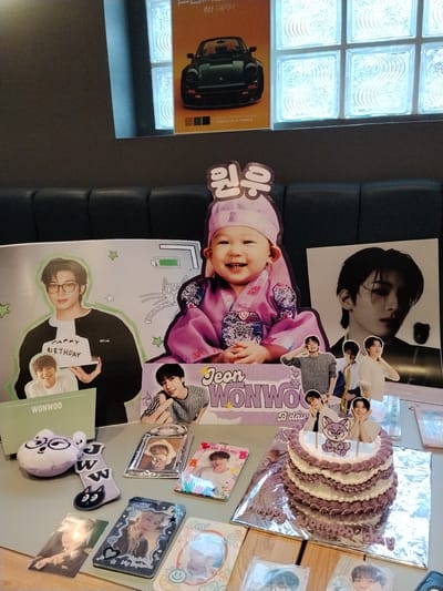The image shows a table setting for a birthday celebration. In the background, a poster of a green Porsche is visible. The table is adorned with various items, including a multi-tiered cake with purple and white frosting, decorated with small photos and a cartoon figure on top. There are also several photo cards, a plush toy, and a keychain. The theme appears to be a K-pop idol's birthday, likely "Jeon Wonwoo" based on the text on the cake and decorations. The overall setting is informal, suggesting a personal celebration. The location in Serang, Indonesia, is not directly evident from the image itself, but this context is provided.