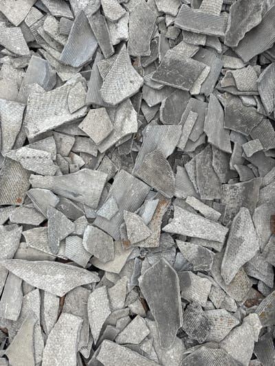 The image is a top-down, close-up view of a pile of broken, gray, corrugated roofing material. The material is fragmented into numerous irregular pieces, ranging in size from small shards to larger slabs. Some pieces have a rough, weathered texture, while others exhibit distinct parallel lines from their corrugated structure. A few flecks of reddish-brown material, possibly rust or dirt, are visible interspersed among the gray fragments. The overall impression is one of debris or demolition waste. There are no people, animals, or identifiable structures in the image. The lighting appears even, suggesting it was taken in daylight, but there are no specific cues to determine the time of day or weather. The location context is provided as Kuybyshev, Russia, but no visual elements within the image itself indicate this specific location. There is no visible text in the image.