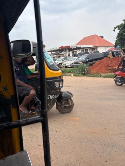 In Ojoto, Nigeria, a yellow and black auto-rickshaw is parked on a dusty, paved road. The driver, a man with dark skin and wearing a black polo shirt and black shorts, is visible from the waist up, looking down. A second man, wearing a green shirt, is seated behind the driver. The auto-rickshaw has the license plate "TMC-4081" visible on its side. In the background, a gas station with a red roof and signage reading "EDEH F. GLOBAL OIL & GAS" is present, with several cars and minivans parked nearby. A large pile of reddish-brown dirt is situated next to the gas station. Further to the right, a red scooter is parked on the road, with a person wearing a black tank top and jeans seated on it. Trees are visible to the far right of the image. The sky is overcast.