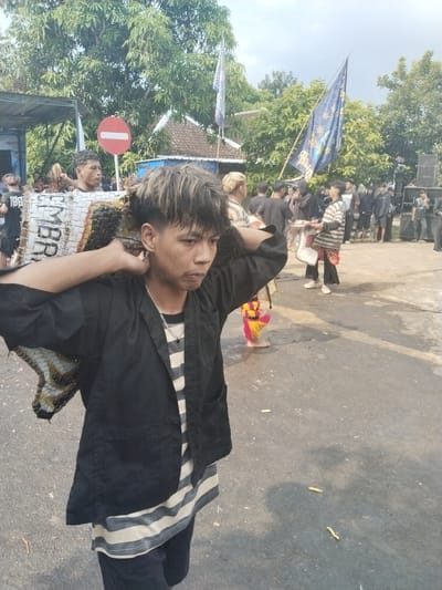 The scene is in Pendem, Indonesia, with a group of people gathered on a paved area or road. The focus is on a young individual with distinctive two-toned hair, wearing a striped shirt, a black jacket, and carrying a large, rectangular woven object on their shoulders. In the background, there are several other people, some wearing similar attire, a flag, and a building. A red and white no-entry sign is visible in the background. There is debris, seemingly spent firecrackers, scattered on the ground. The overall impression is of a cultural event or celebration.