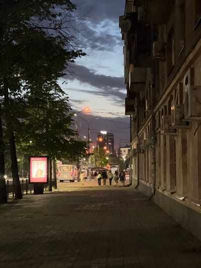 The image shows a street scene in the city of посёлок 1-го отделения совхоза "Масловский", Russia at dusk or dawn. On the left side of the image, trees with dense foliage are visible, obscuring the sky above. Buildings with intricate architectural details are overhead on both sides of the street. People are walking along the sidewalk on the right, and a bus is approaching. There is a billboard with a red sign on the right side of the street. Streetlights illuminate the scene.